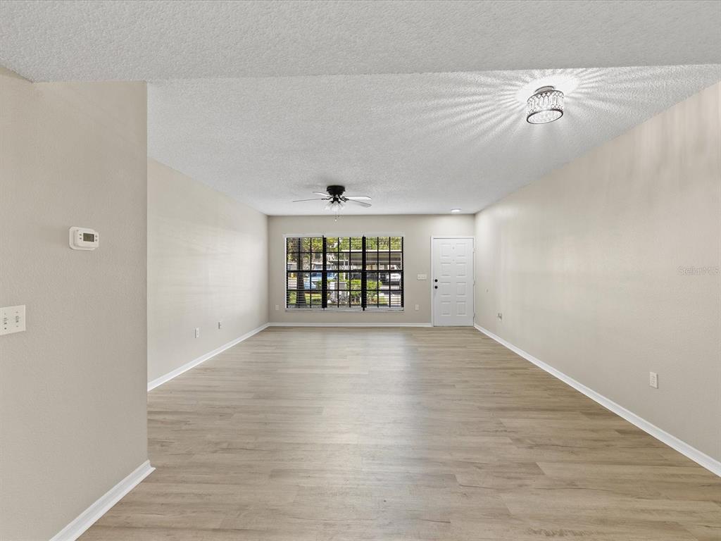 An empty living room features light wood-look flooring, beige walls, a ceiling fan, and a window — 3358 MERMOOR DRIVE UNIT 102, PALM HARBOR, FL