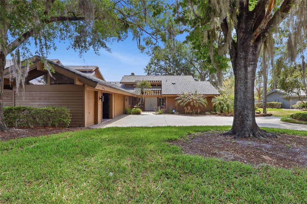 A brown wood-sided house with a large driveway and mature trees under a blue sky — 29922 BAYWOOD LANE, WESLEY CHAPEL, FL
