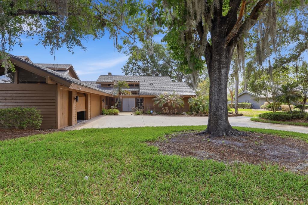 A brown wood-sided house with a multi-car garage and a large tree in the yard — 29922 BAYWOOD LANE, WESLEY CHAPEL, FL