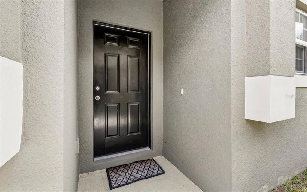 A black front door is set into a gray stucco exterior wall with a doormat — 18307 TURNING LEAF CIRCLE, LAND O LAKES, FL