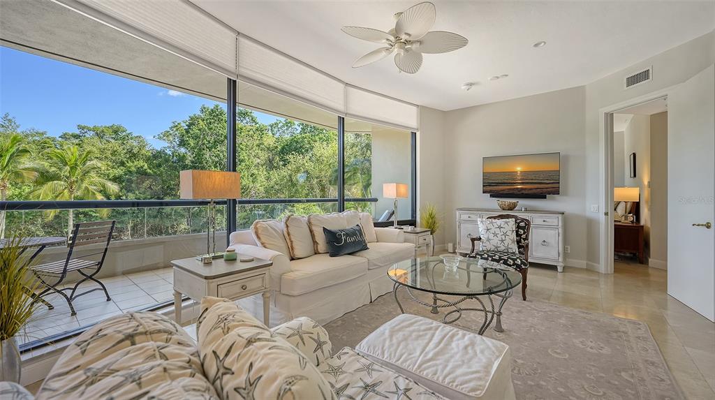 A living room with a white sofa, glass coffee table, and large windows overlooking trees — 1241 GULF OF MEXICO DRIVE UNIT 111, LONGBOAT KEY, FL