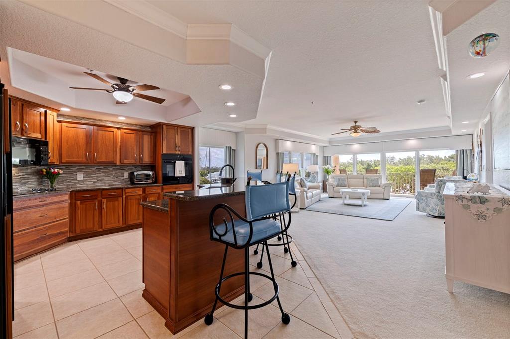 An open-concept kitchen with wood cabinets and a breakfast bar overlooks a carpeted living area — 1645 PINELLAS BAYWAY S UNIT A1, TIERRA VERDE, FL
