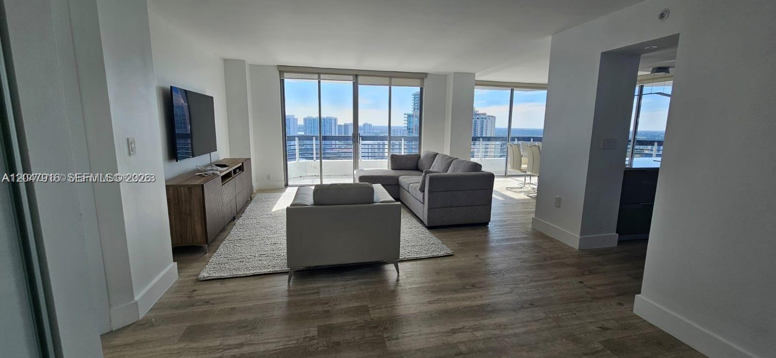 A living room features light wood flooring, a grey sectional sofa, and floor-to-ceiling windows — AVENTURA, FL
