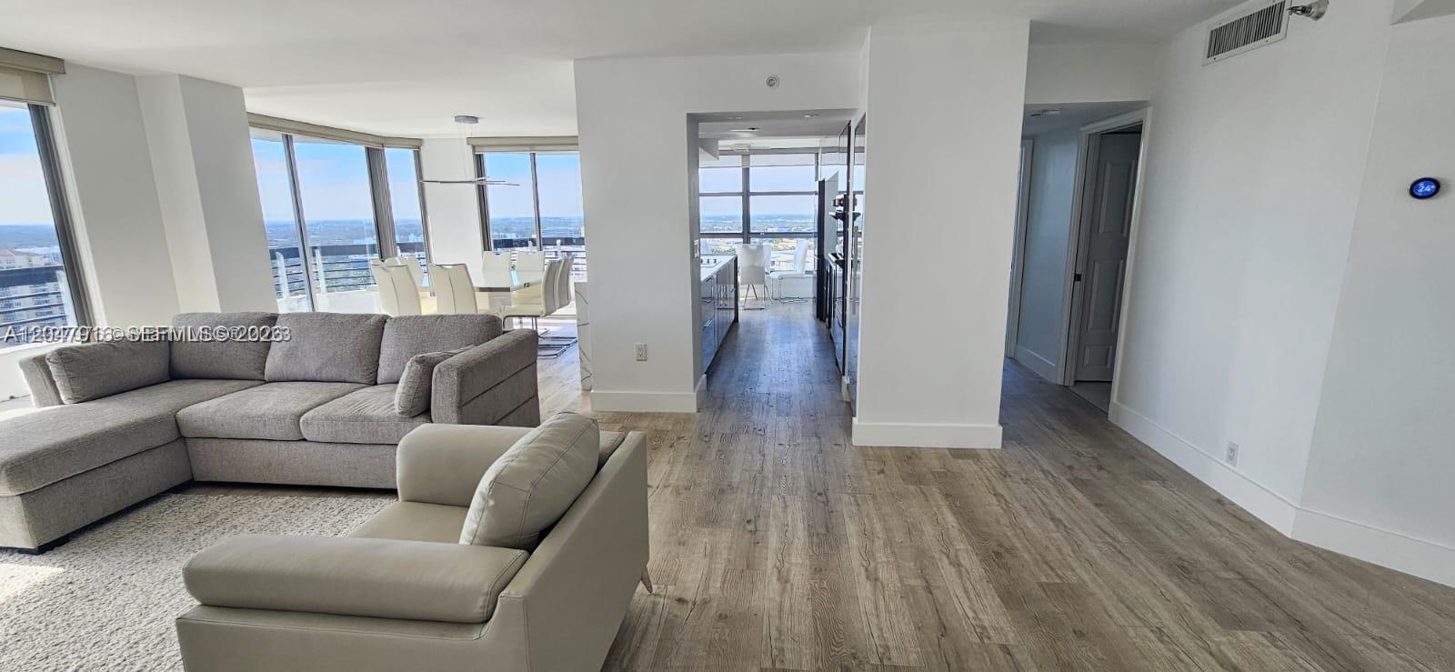 A modern living room features light wood flooring, a grey sectional sofa, and floor-to-ceiling windows — AVENTURA, FL