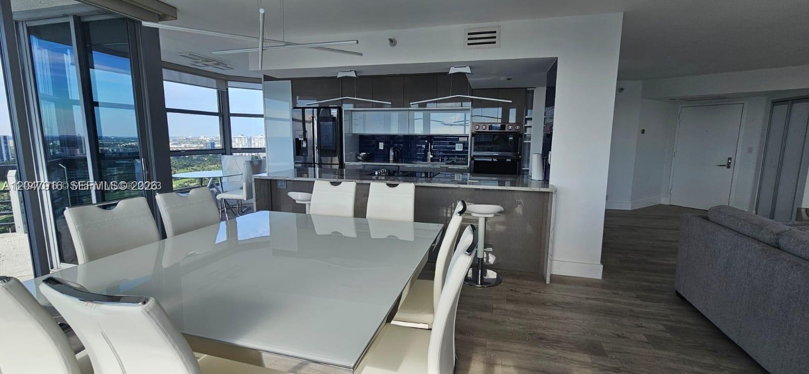 A modern dining area and kitchen feature white chairs, a glass table, and city views — AVENTURA, FL