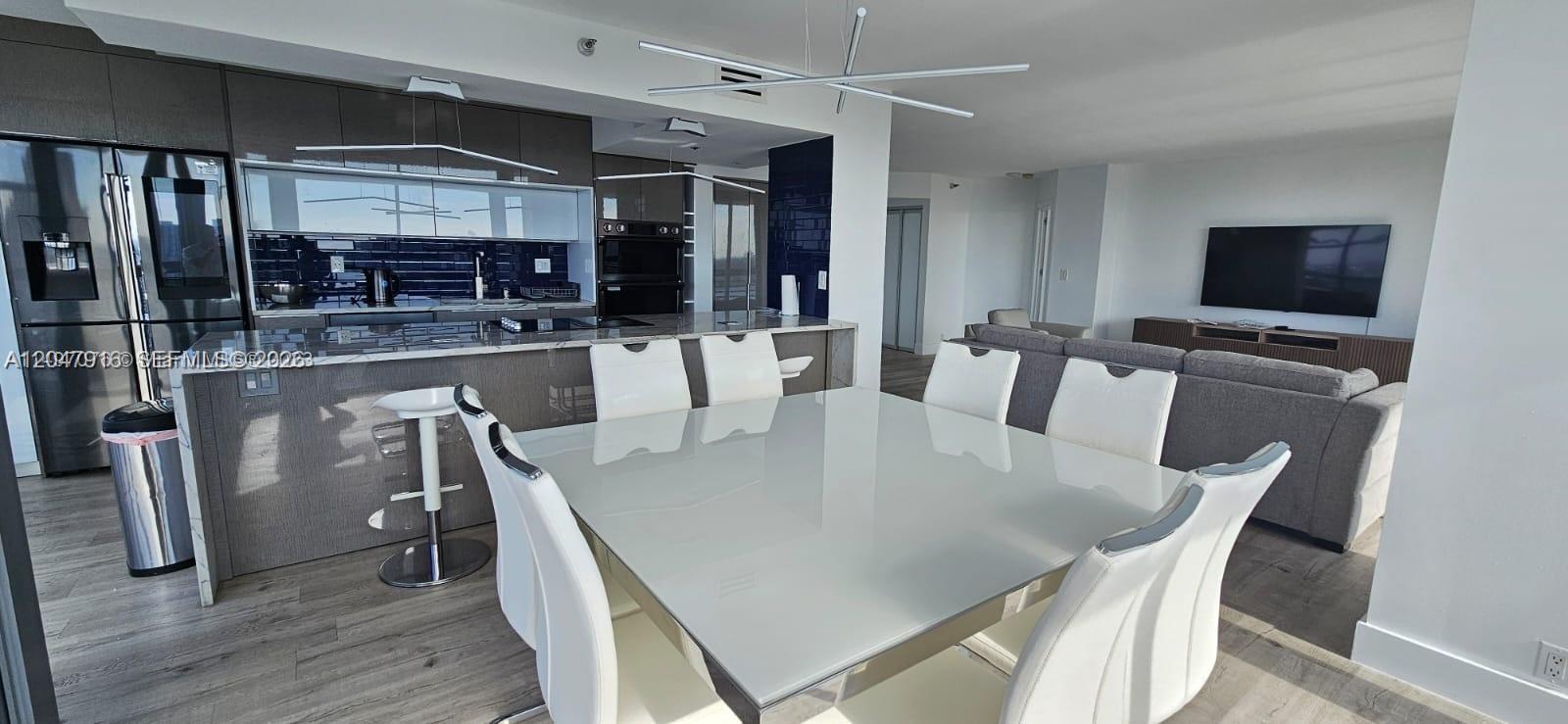A modern open-concept kitchen and dining area features white chairs, a glass table, and cabinetry — AVENTURA, FL