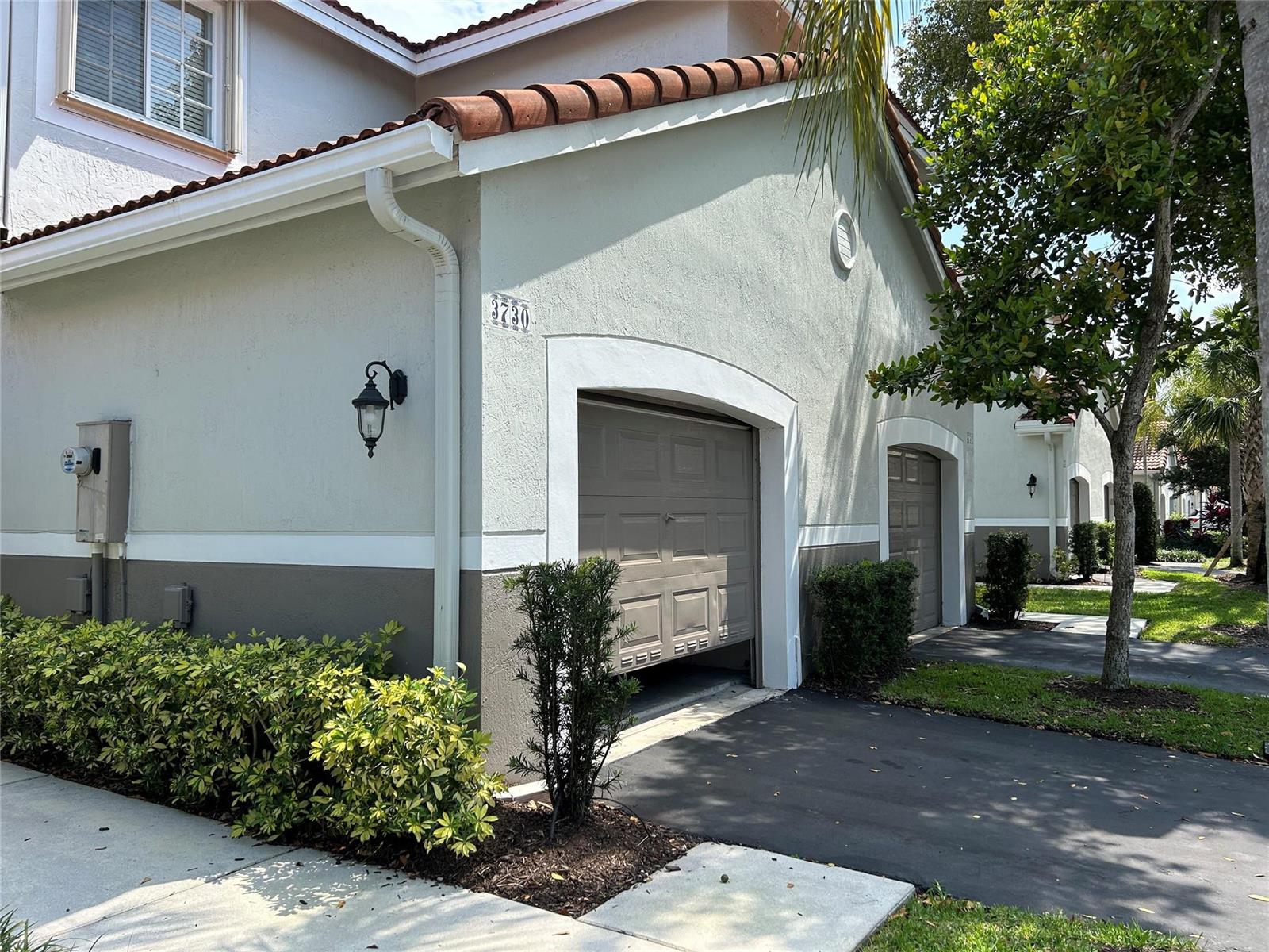 A view of a tan stucco garage exterior with a partially open door and landscaping — 3730 San Simeon Circle, Weston FL 33331, WESTON, FL