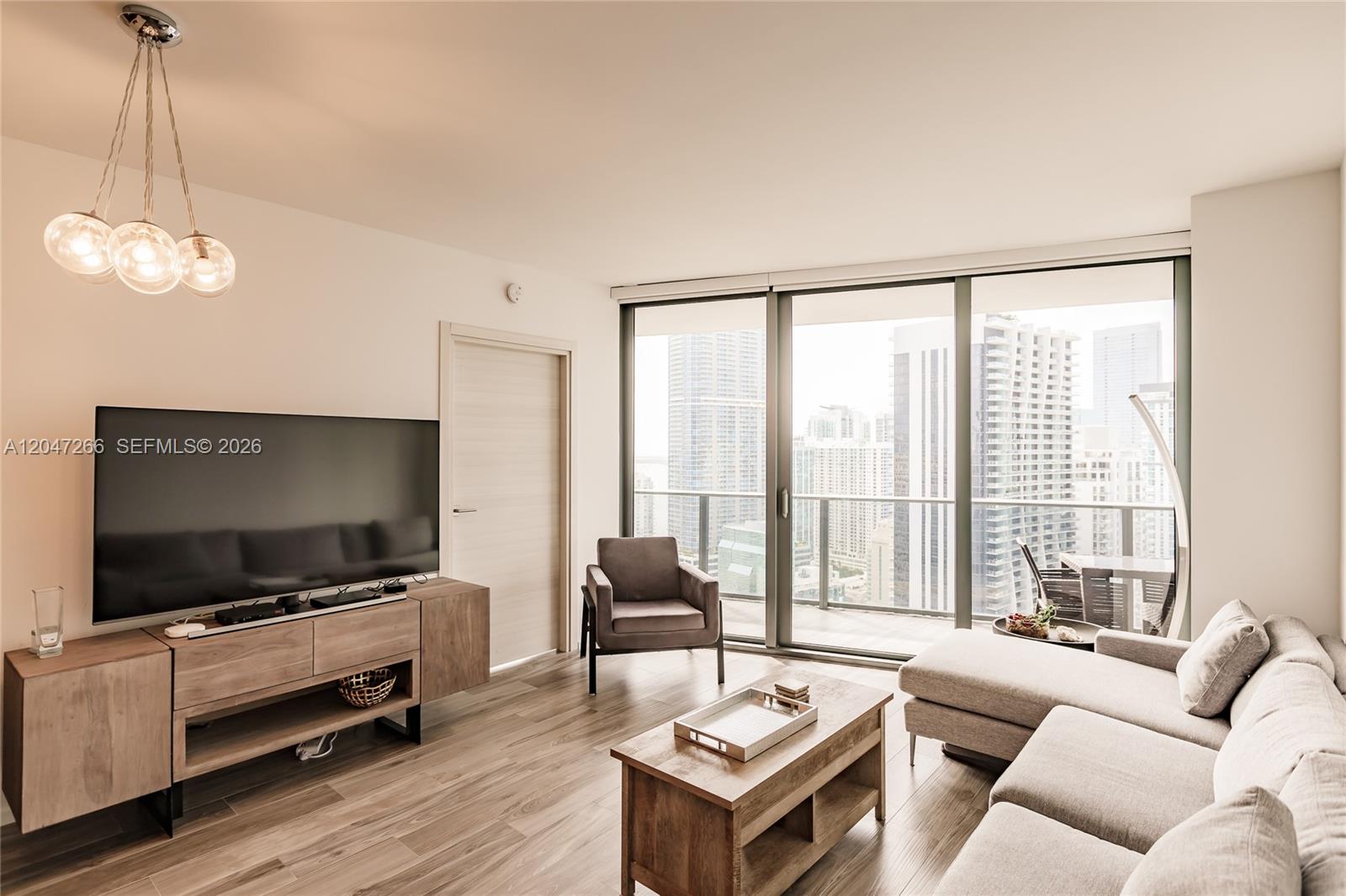 A modern living room features a television, sectional sofa, and floor-to-ceiling windows overlooking city buildings — 801 S Miami Ave # 3703, Miami FL 33130, MIAMI, FL