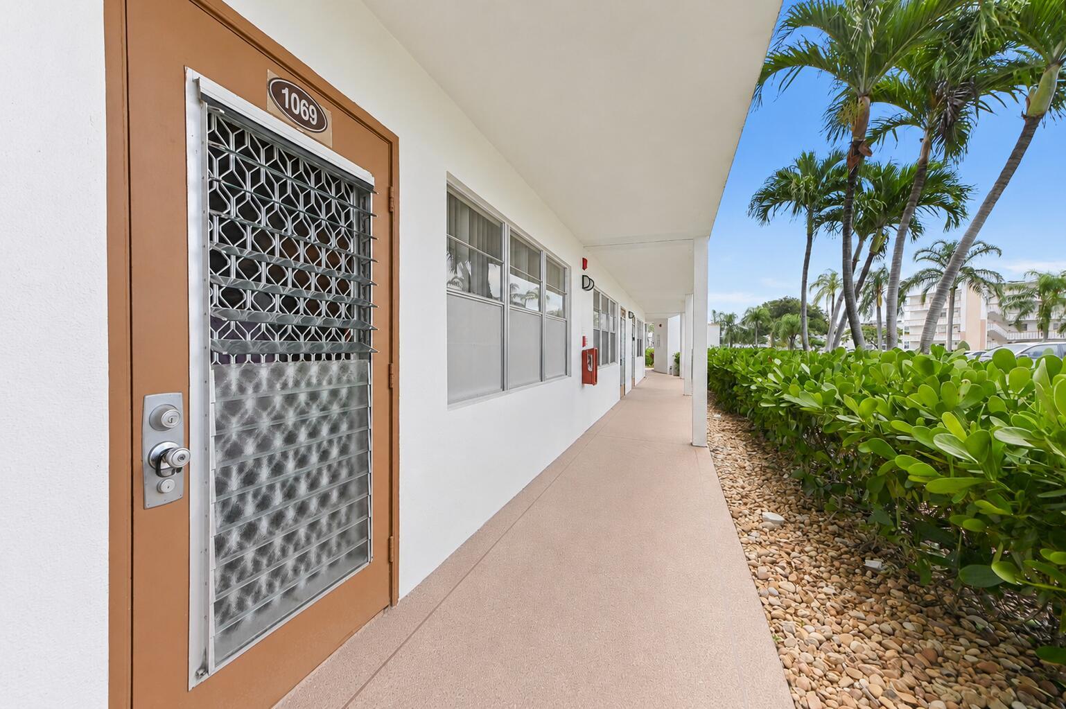 An exterior walkway leads past a brown door with a security screen and palm trees — 1069 Cornwall D, Boca Raton FL 33434, BOCA RATON, FL