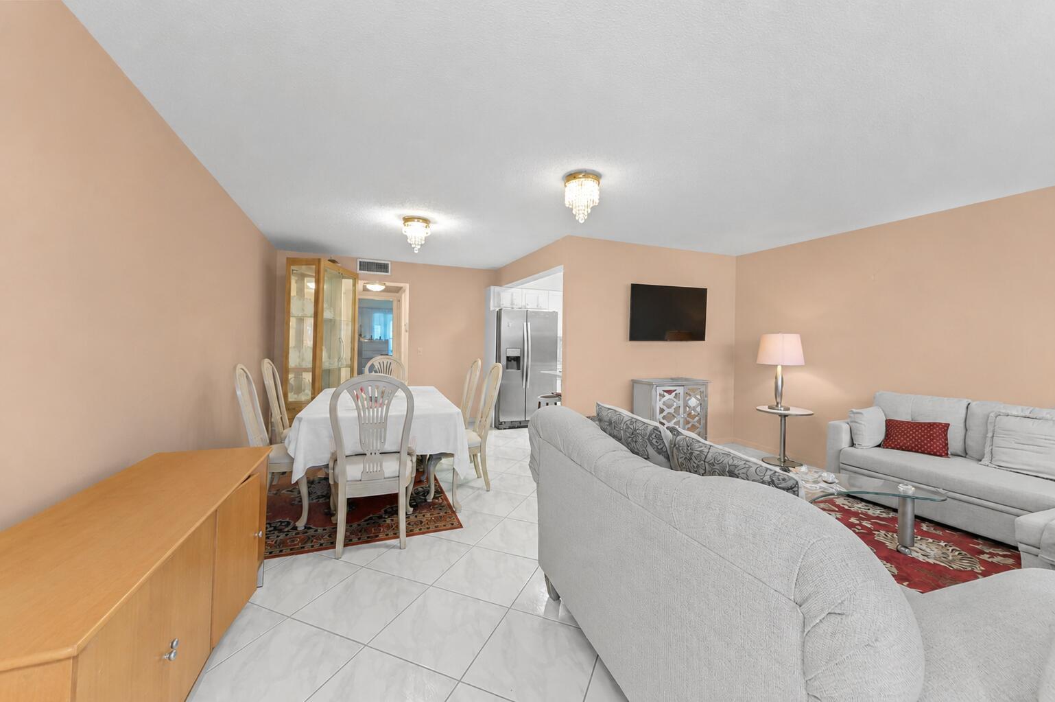An open-concept living and dining area features light-colored tile flooring, peach walls, and chandeliers — 1069 Cornwall D, Boca Raton FL 33434, BOCA RATON, FL