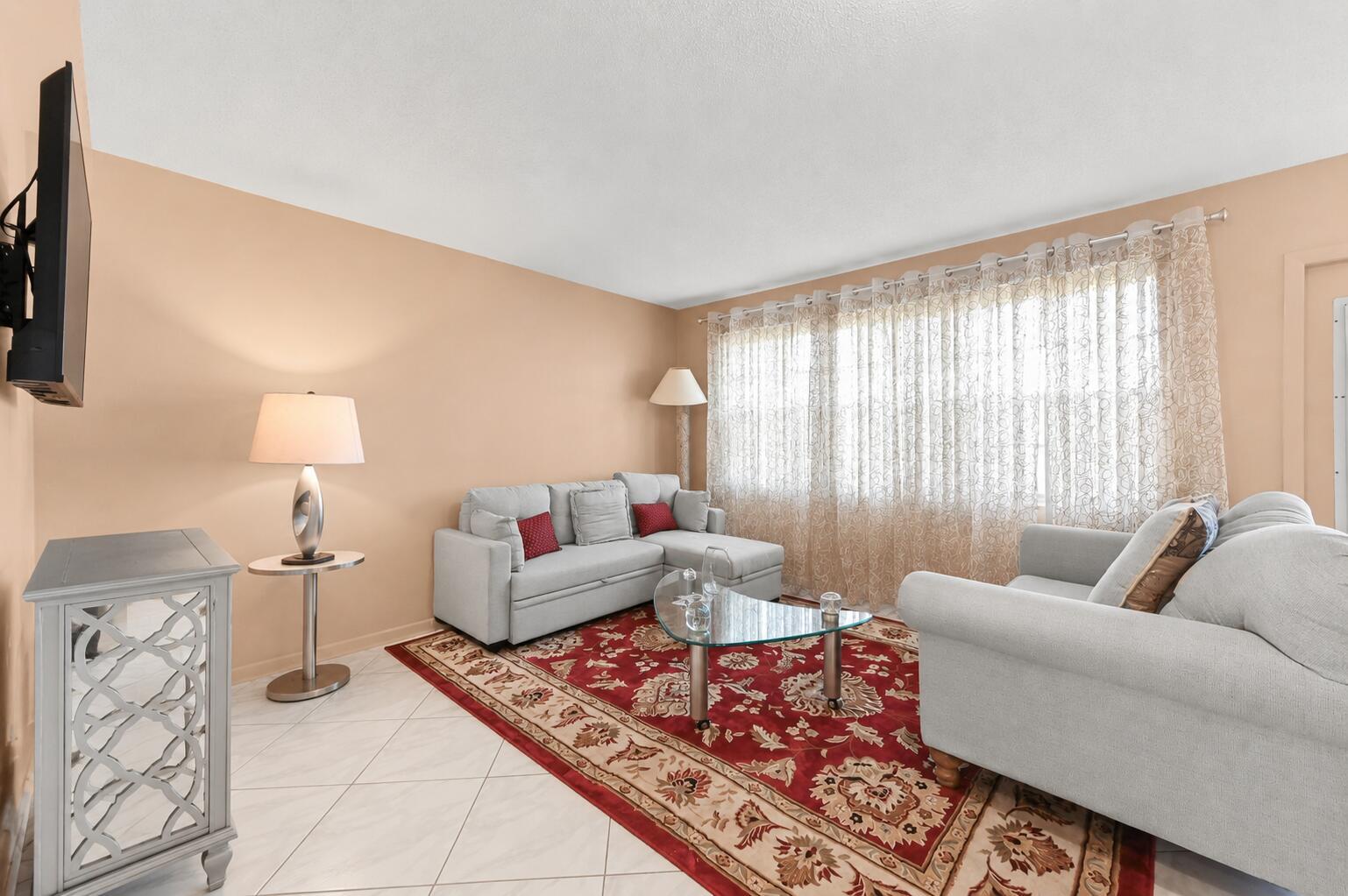 A living room features light-colored sofas, a red patterned area rug, and sheer window curtains — 1069 Cornwall D, Boca Raton FL 33434, BOCA RATON, FL
