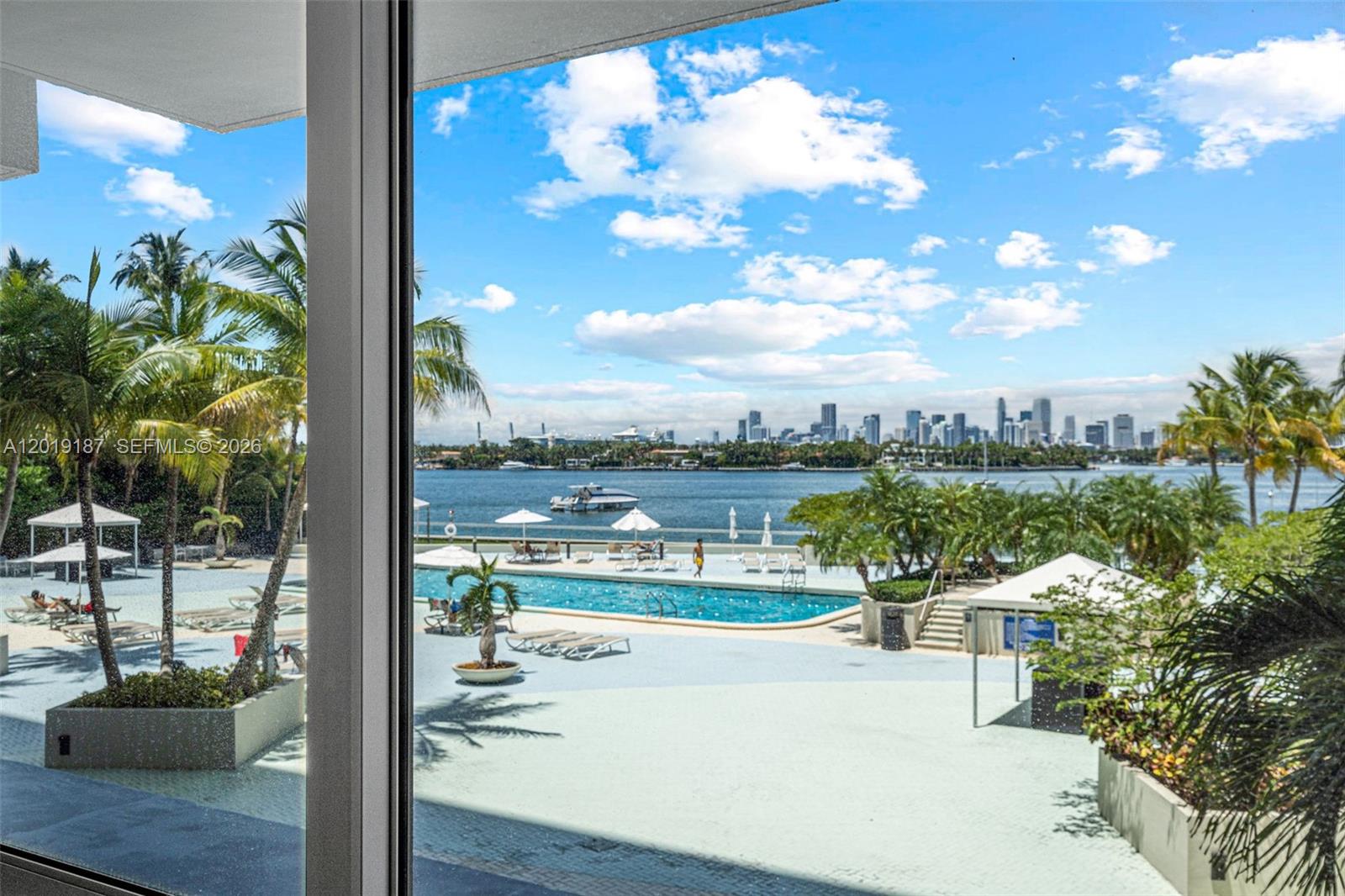 A view from a window shows a swimming pool, palm trees, and a distant city skyline — 1200 West Ave # 216, Miami Beach FL 33139, MIAMI BEACH, FL