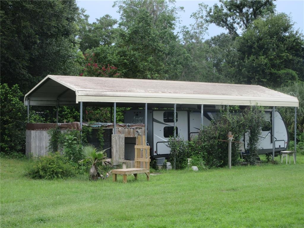 A travel trailer is parked under a large metal carport in a grassy, wooded yard — 16000 N HWY 329, REDDICK, FL