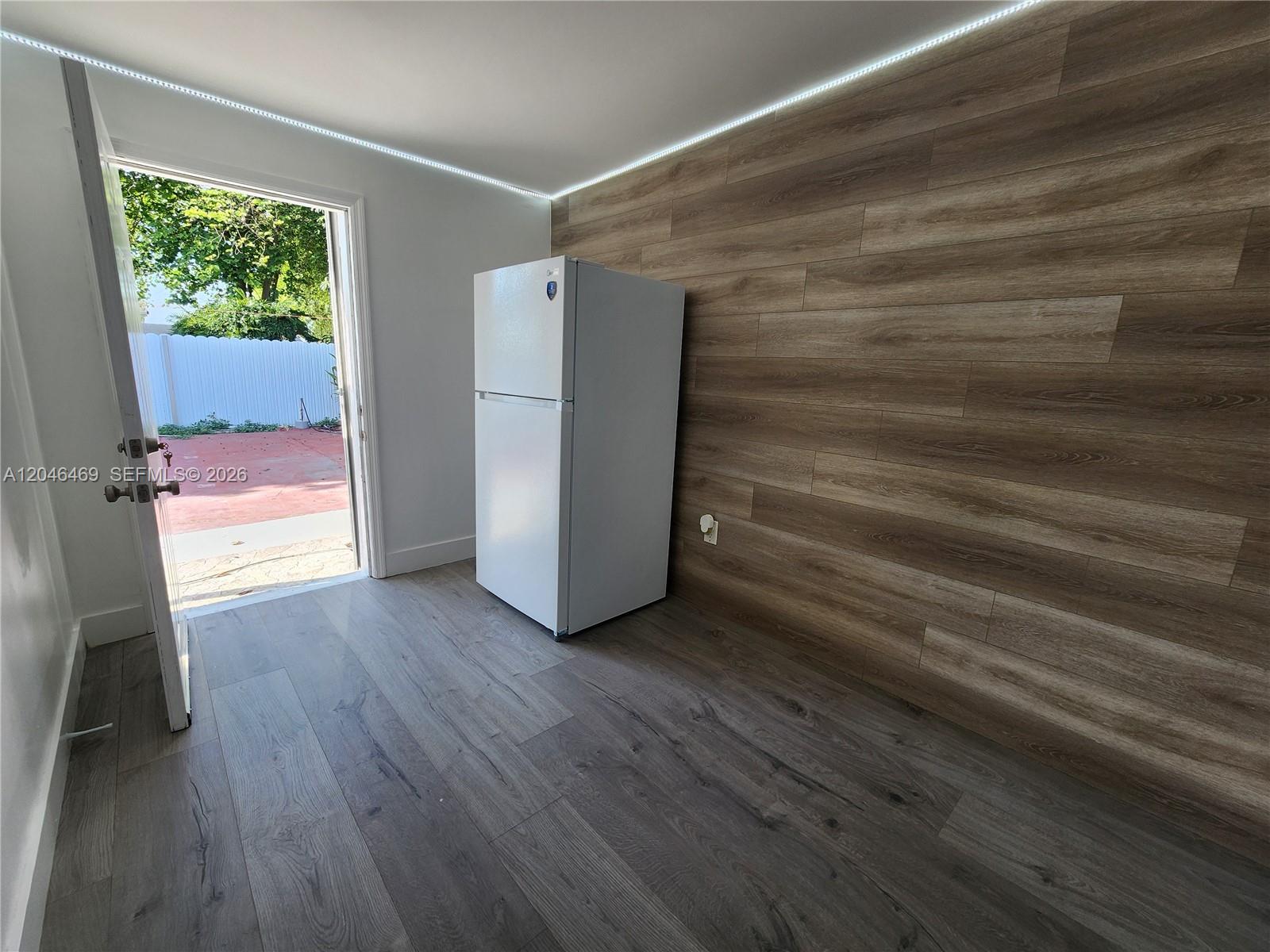 A room with wood-look flooring, a wood-paneled accent wall, a white refrigerator, and an open door — 11461 SW 197th St # A, Miami FL 33157, MIAMI, FL