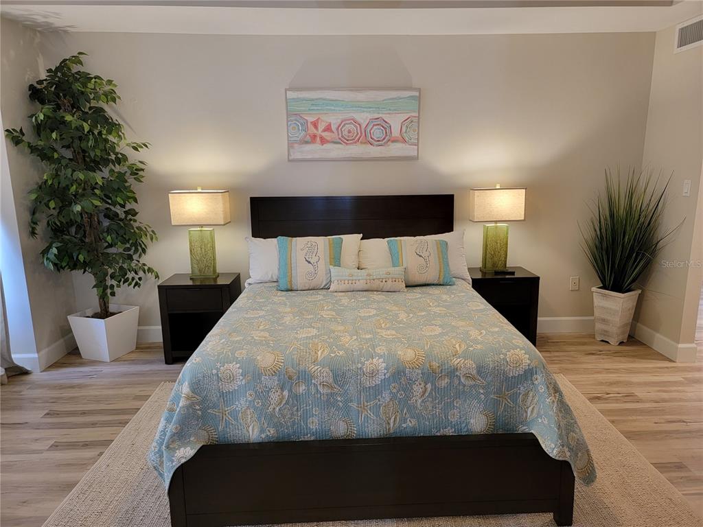 A bedroom features a dark wood bed frame, coastal-themed bedding, two lamps, and two potted plants — 7650 BAYSHORE DRIVE UNIT 302, TREASURE ISLAND, FL