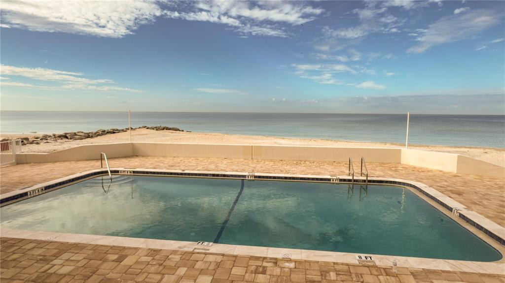 A rectangular swimming pool sits on a paved patio overlooking a sandy beach and ocean — 1900 BENJAMIN FRANKLIN DRIVE UNIT 201BER, SARASOTA, FL