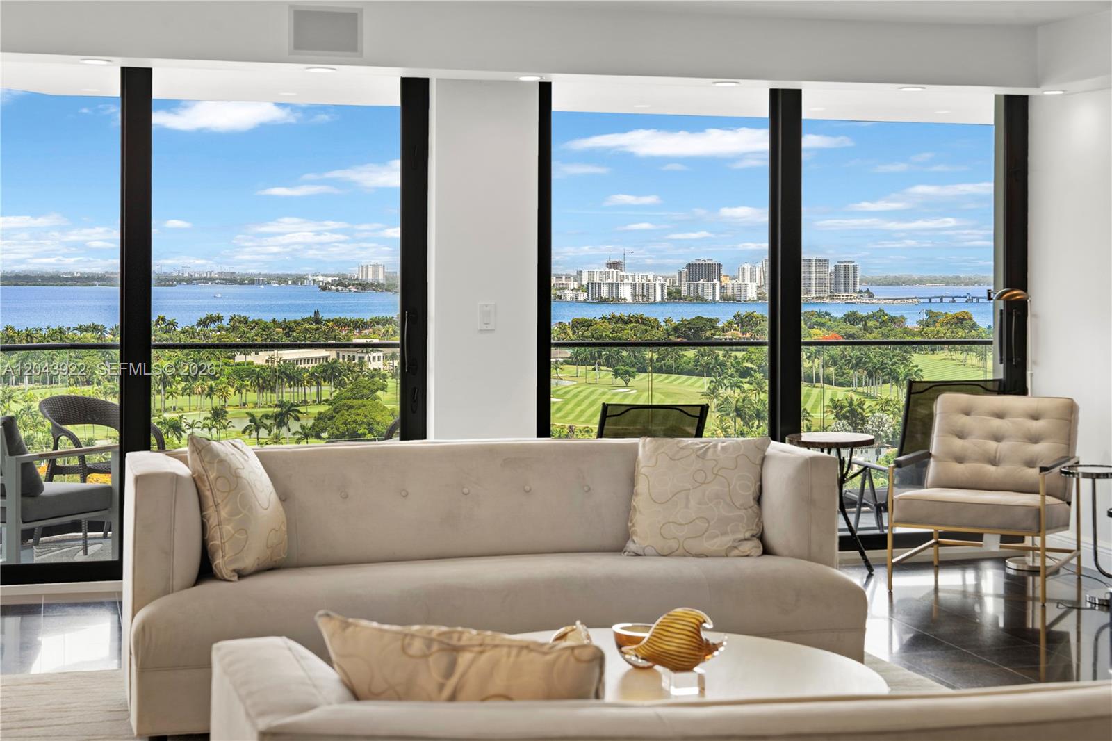 A living room features a beige sofa, tufted accent chair, and floor-to-ceiling windows overlooking a golf course — 5500 Collins Ave # 1902, Miami Beach FL 33140, MIAMI BEACH, FL