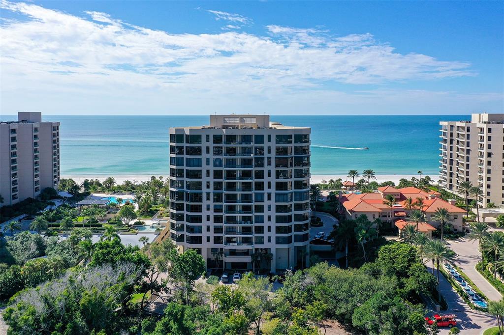 An aerial view shows a multi-story residential building situated on a beach with lush landscaping — 1241 GULF OF MEXICO DRIVE UNIT 303, LONGBOAT KEY, FL