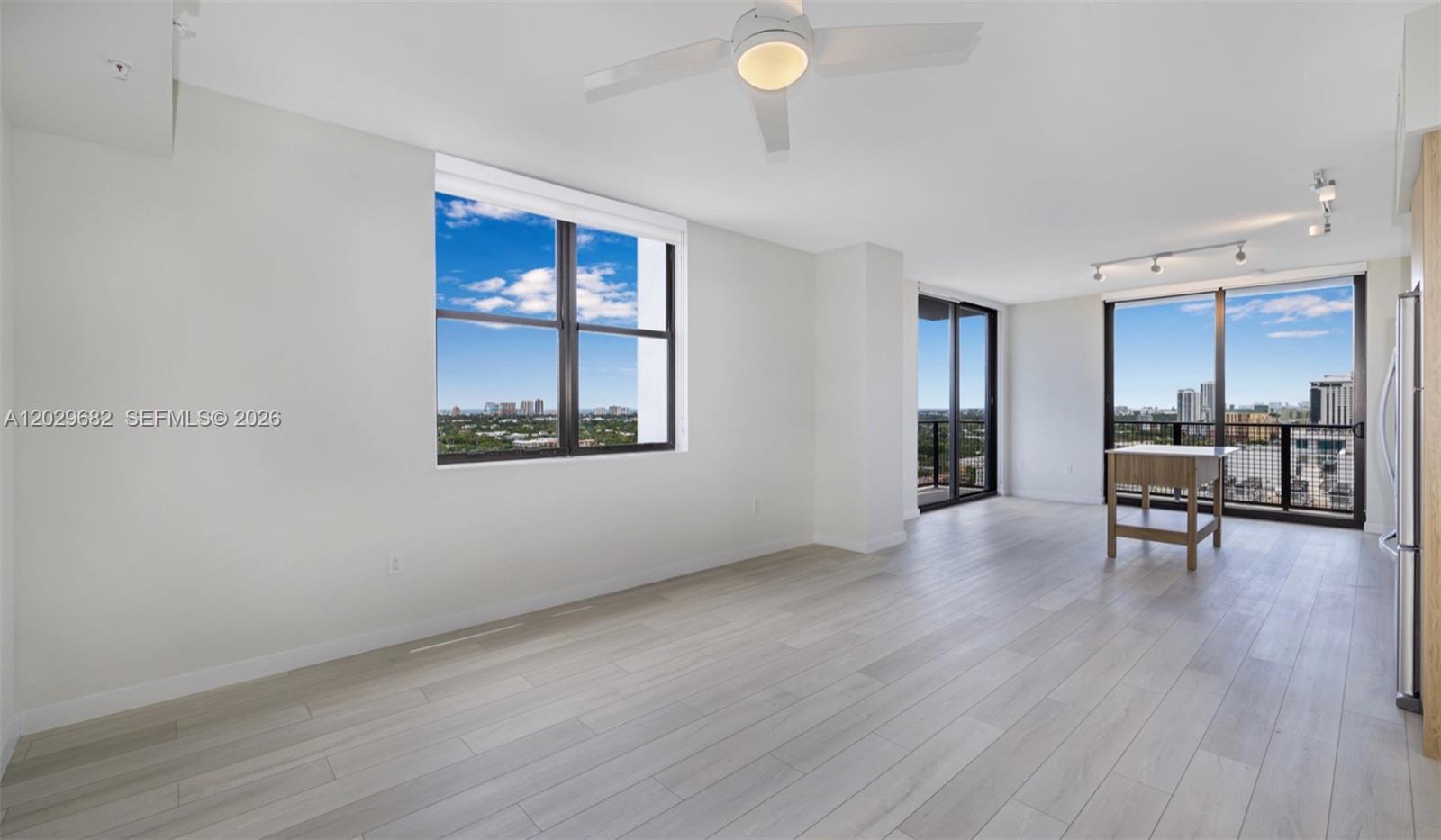 An empty living area features light wood-look flooring, a ceiling fan, and floor-to-ceiling windows — FORT LAUDERDALE, FL