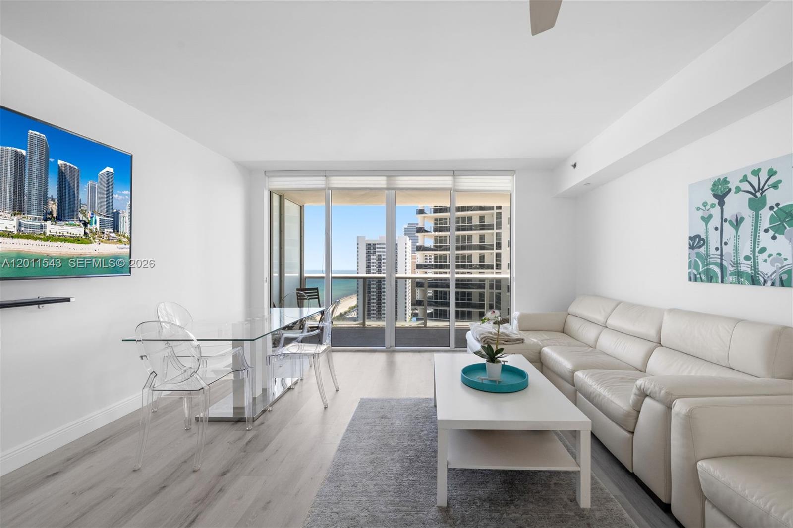 A bright living and dining room features a white sofa, glass table, and balcony view — HALLANDALE BEACH, FL
