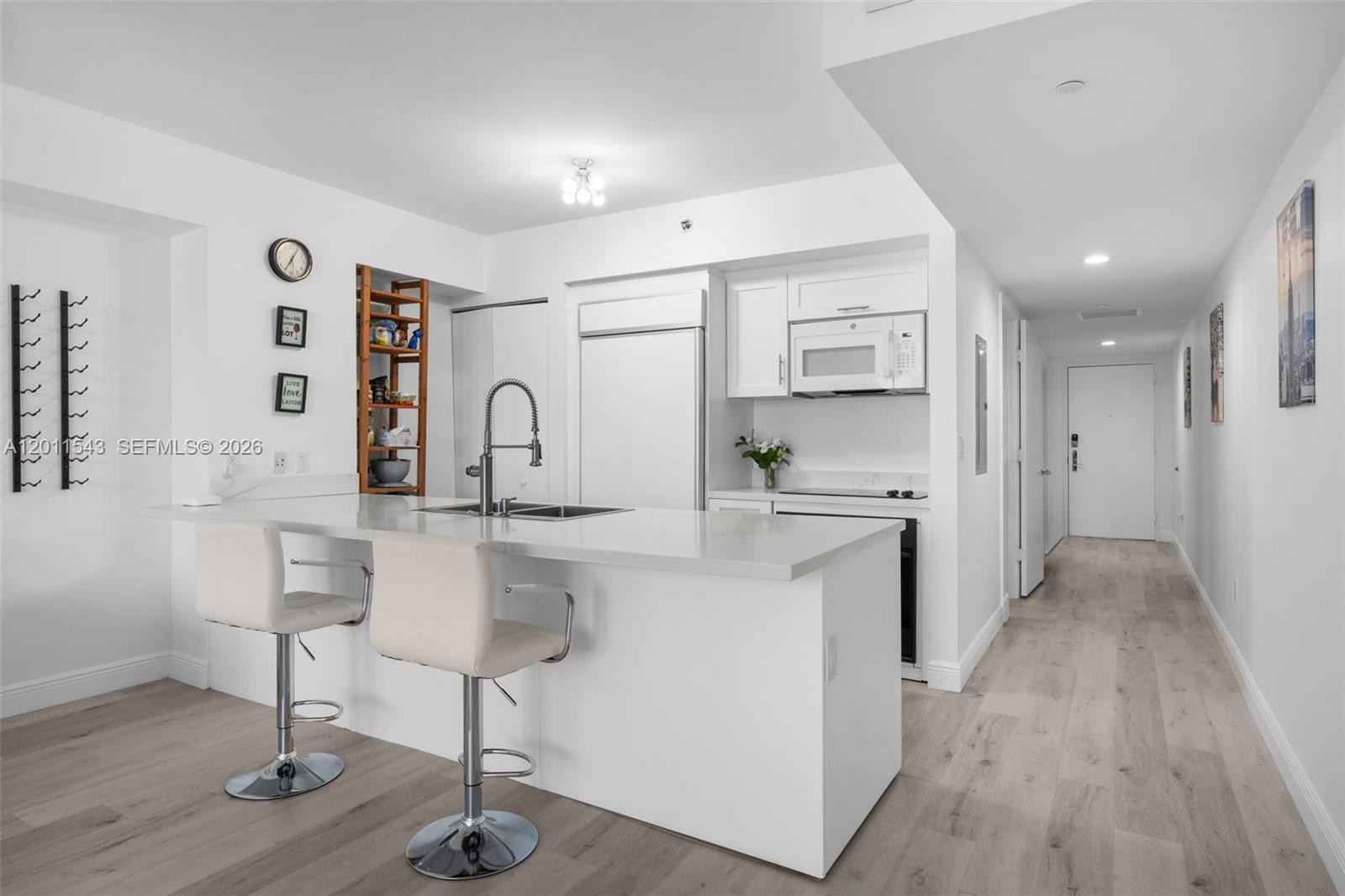 A modern white kitchen features a breakfast bar with two stools and light wood flooring — HALLANDALE BEACH, FL