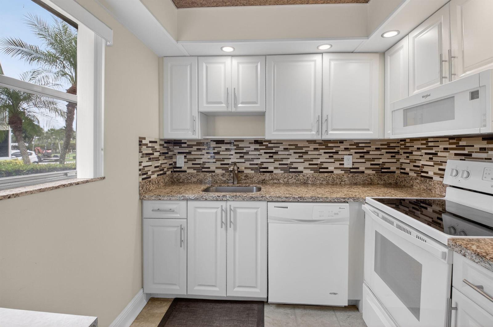 A kitchen features white cabinets, granite countertops, a mosaic tile backsplash, and a white dishwasher — 204 Flanders E, Delray Beach FL 33484, DELRAY BEACH, FL