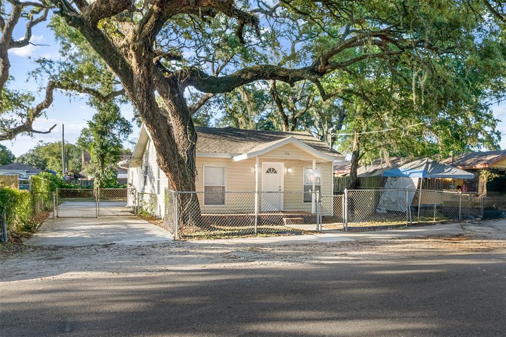 A single-story house with light siding stands behind a chain-link fence under large trees — 2116 E ELLICOTT STREET, TAMPA, FL