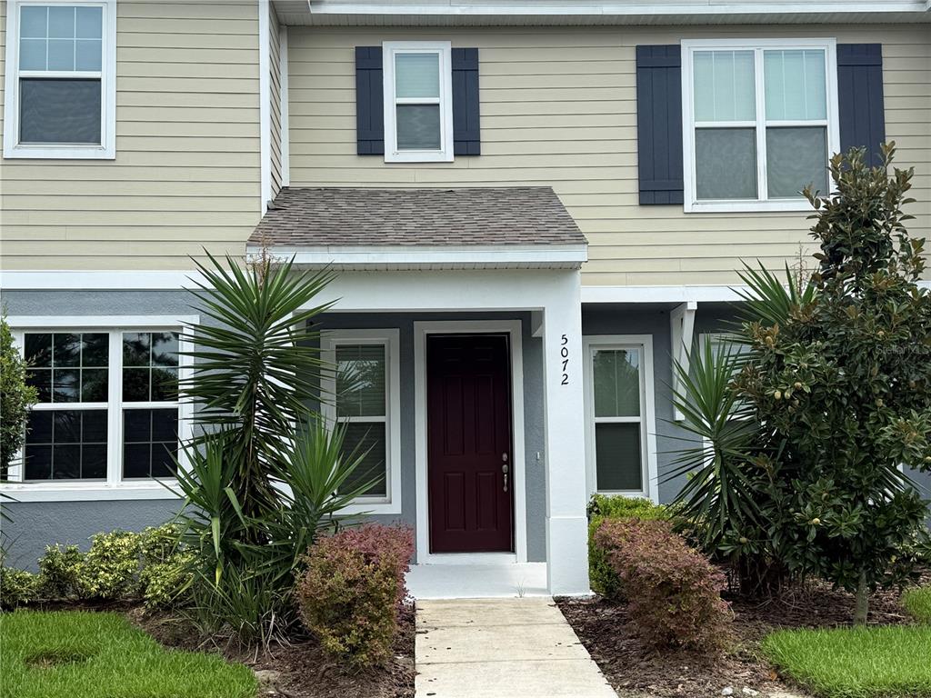 A two-story townhouse exterior features tan siding, a dark red front door, and landscaping — 5072 AVALON PARK BOULEVARD, WESLEY CHAPEL, FL