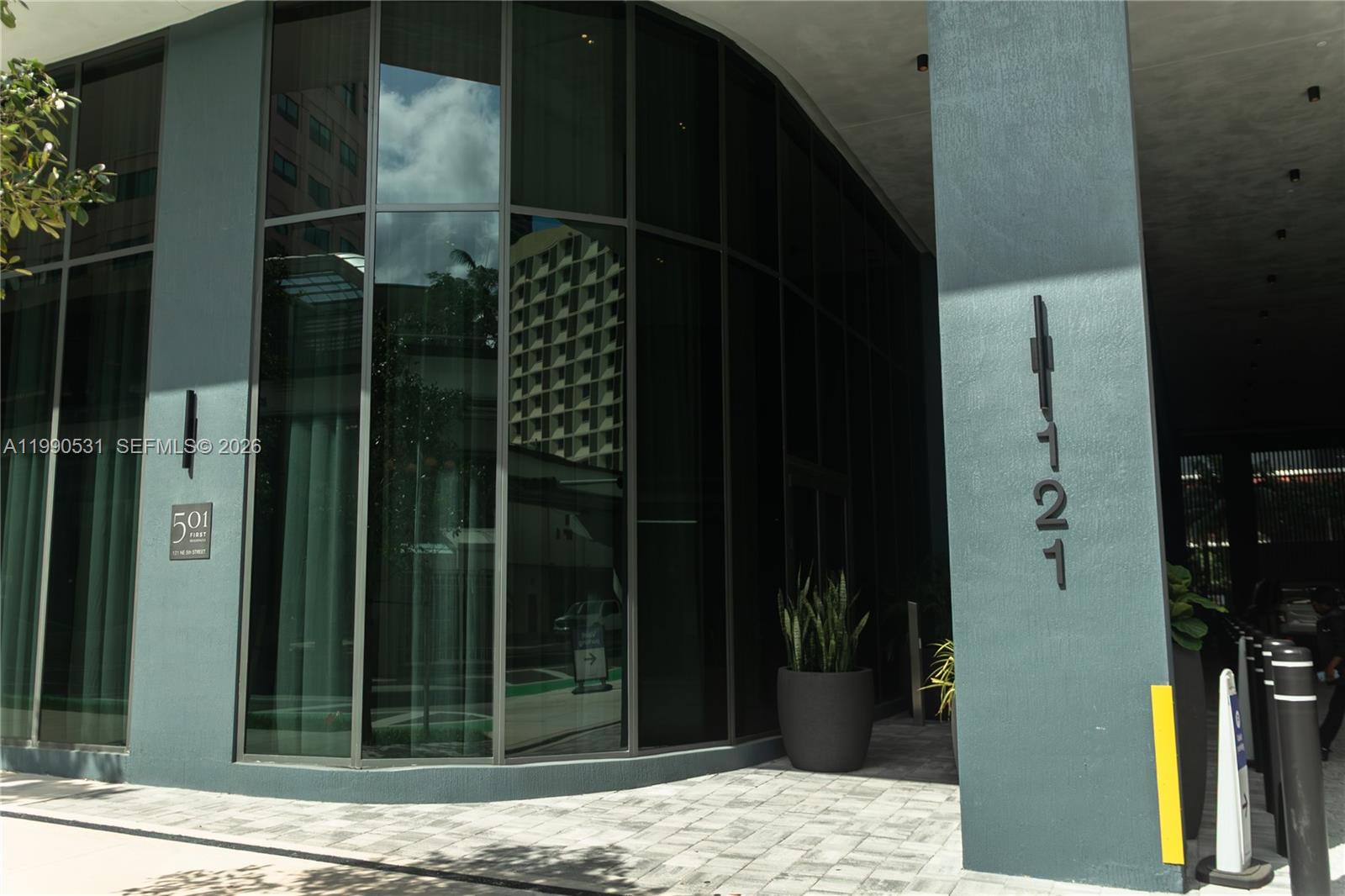 A modern glass building entrance features double doors, dark planters, and black bollards — 121 NE 5th St # 2611, Miami FL 33132, MIAMI, FL