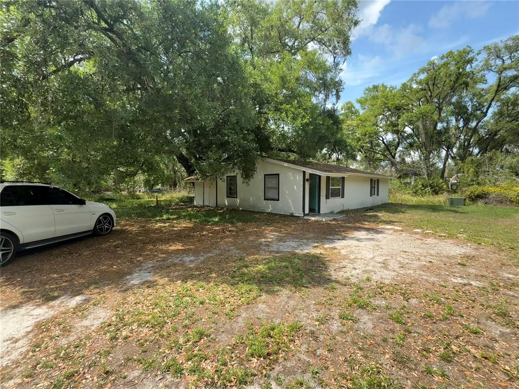 A single-story house with a white exterior sits on a dirt lot under large trees — 3424 MAZE LANE, BRANDON, FL