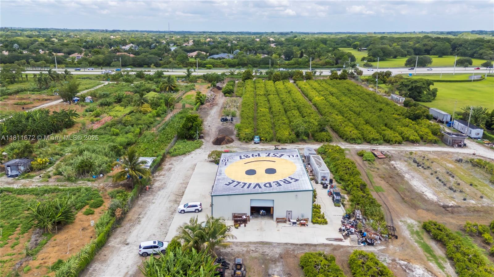 An aerial view shows a warehouse with a smiley face roof mural surrounded by farmland — 4077 SW 121st Ter, Davie FL 33330, DAVIE, FL