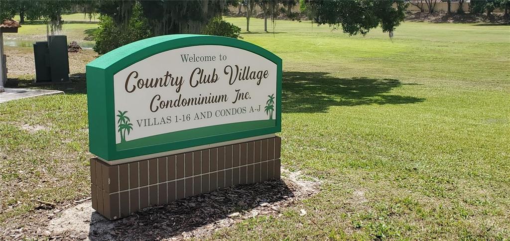 A green and white sign for Country Club Village Condominium stands on a grassy lawn — 10202 VILLAGE DRIVE UNIT 22, LAKE WALES, FL