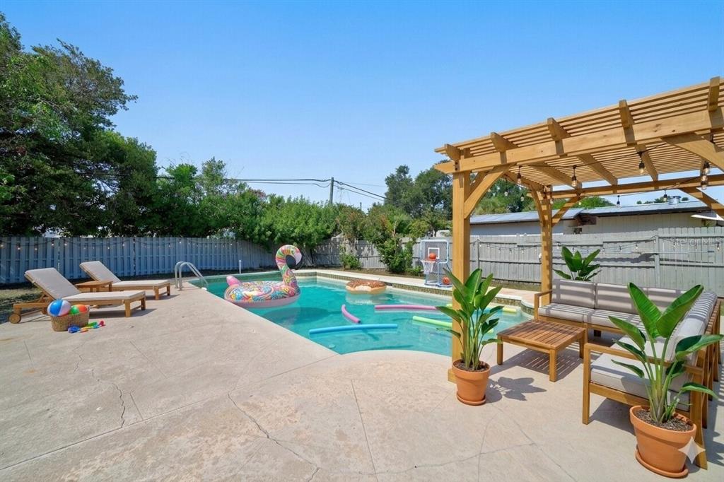 A backyard swimming pool features a wooden pergola, lounge chairs, and a colorful flamingo float — 10760 62ND AVENUE, SEMINOLE, FL
