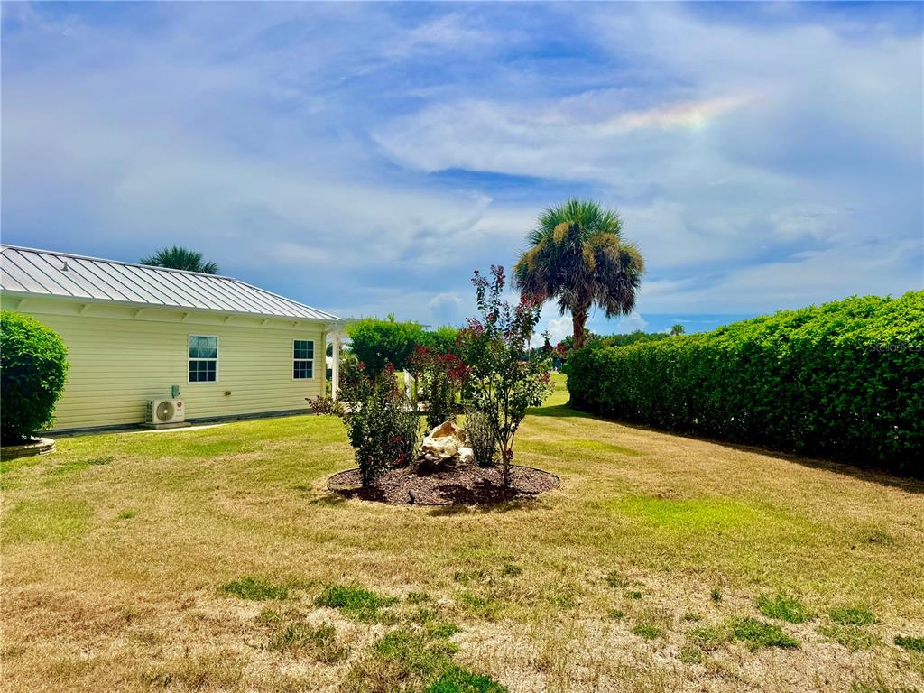 A backyard features a light yellow house, a central landscaped garden bed, and a tall hedge — 9444 SE 49TH TERRACE, WEBSTER, FL