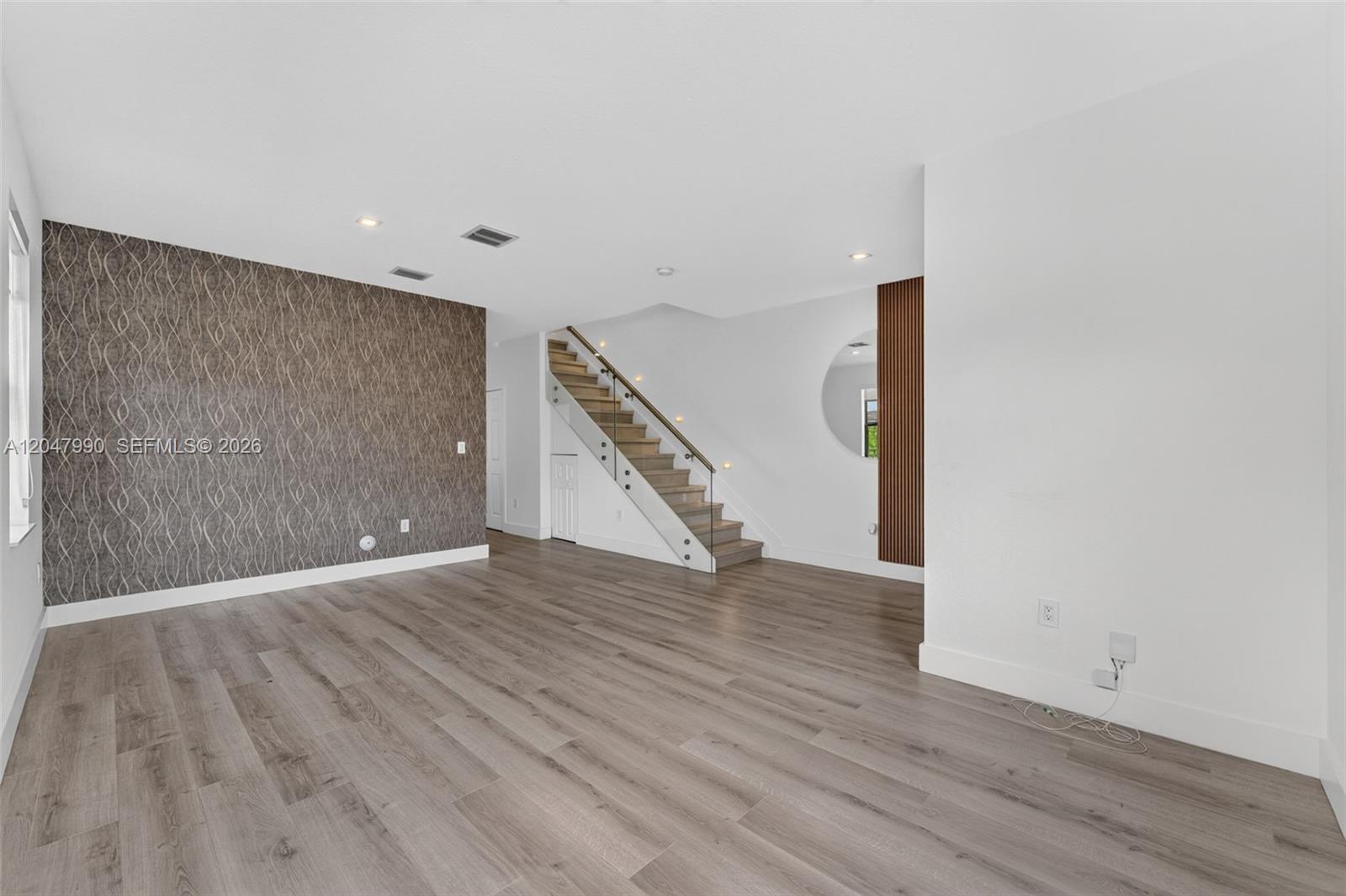 An empty room features wood-look flooring, a patterned accent wall, and a glass-railed staircase — 10908 W 32nd Ln, Hialeah FL 33018, HIALEAH, FL