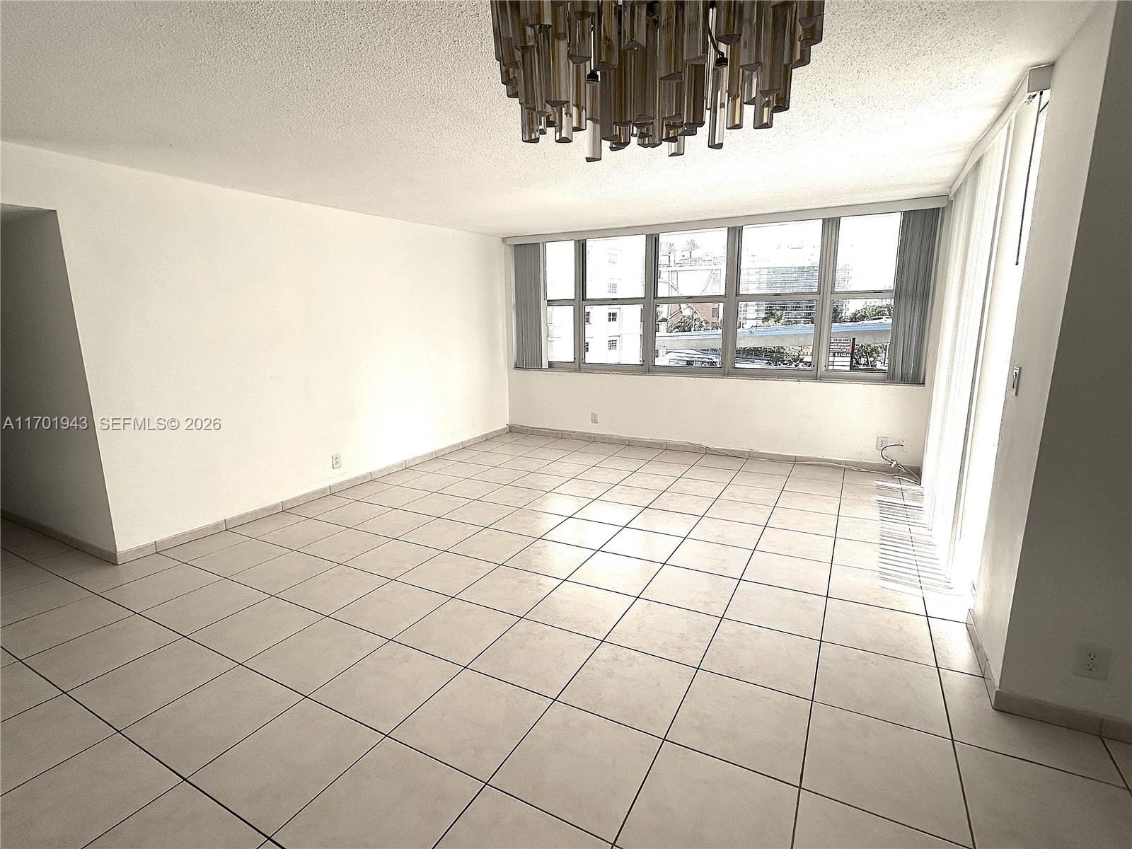 An empty room features white walls, light-colored tile flooring, and a large modern chandelier — 1825 S Ocean Dr # 401, Hallandale Beach FL 33009, HALLANDALE BEACH, FL