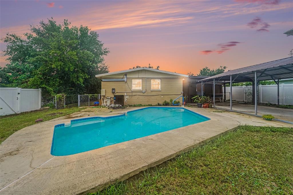 A backyard features a swimming pool, a tan house, and a metal carport at sunset — 4224 E REGNAS AVENUE, TAMPA, FL