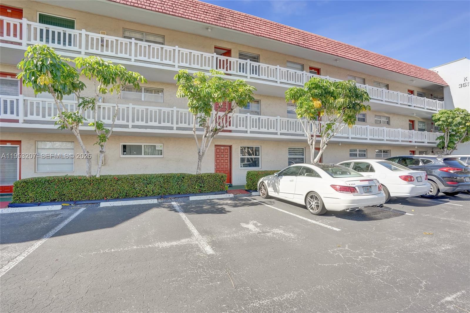 A three-story apartment building features white balconies, red doors, and a parking lot in front — 3511 E Inverrary Dr # 107, Lauderhill FL 33319, LAUDERHILL, FL