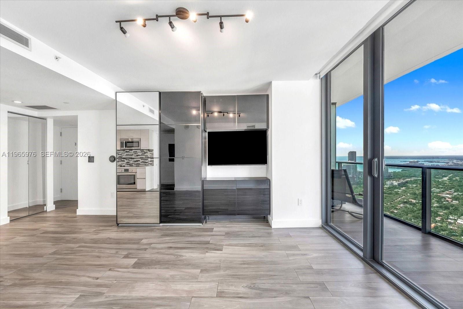 A modern living room features wood-look tile floors, a mirrored storage unit, and a balcony view — 1300 S Miami Ave # 4807, Miami FL 33130, MIAMI, FL