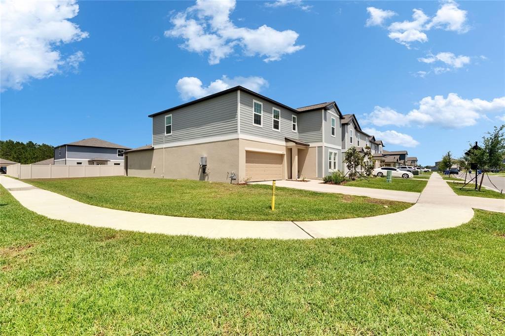 A two-story suburban townhouse with a tan garage and gray siding sits on a corner lot — 34780 RAINVILLE LOOP, ZEPHYRHILLS, FL