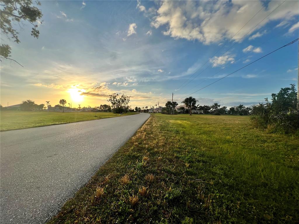 A paved road runs through a grassy field under a sunset sky with scattered clouds — 24268 PEPPERCORN ROAD, PUNTA GORDA, FL