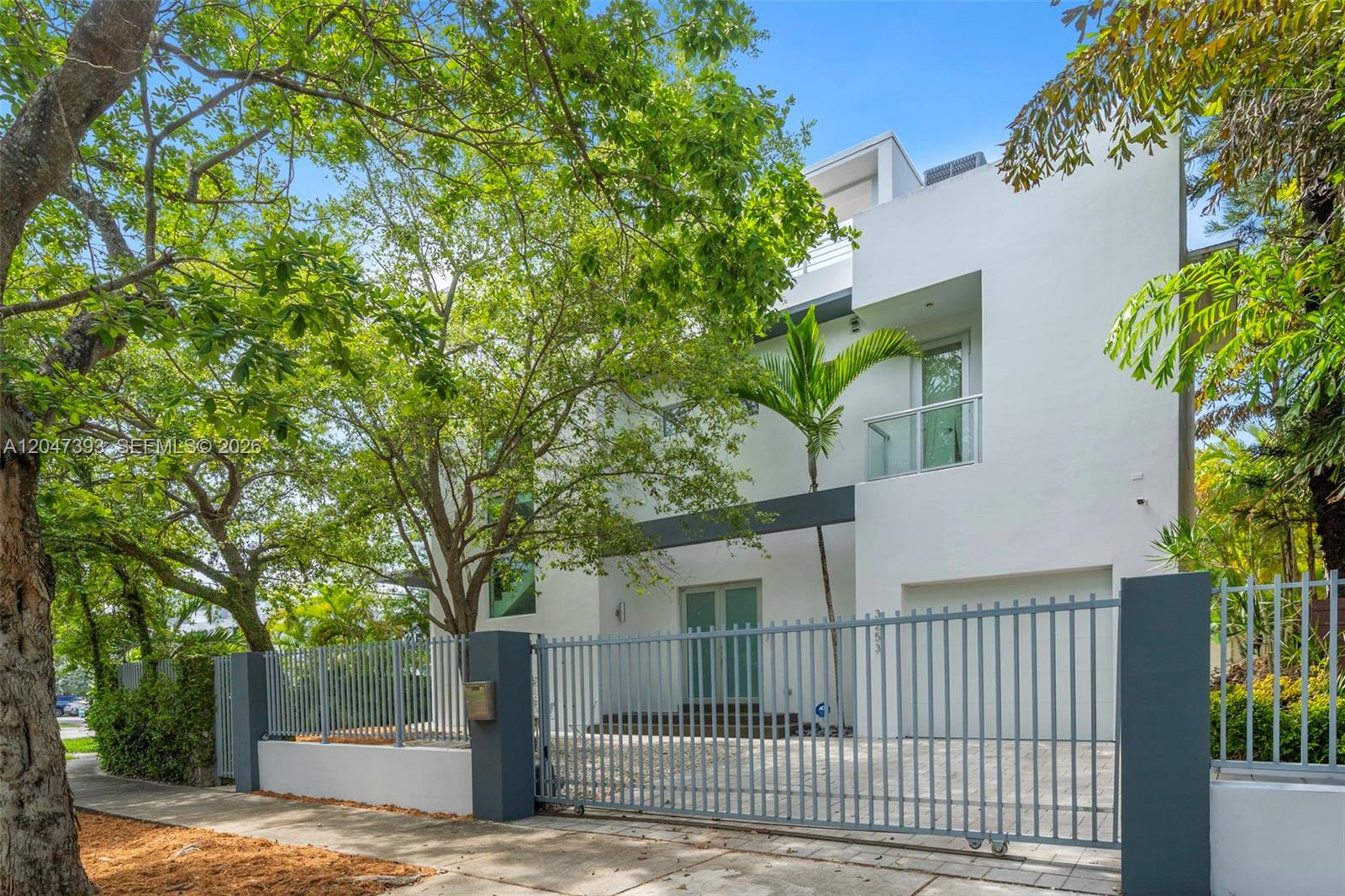 A modern white three-story house is viewed behind a gray metal gate and fence — 3253 Mcdonald St, Miami FL 33133, MIAMI, FL