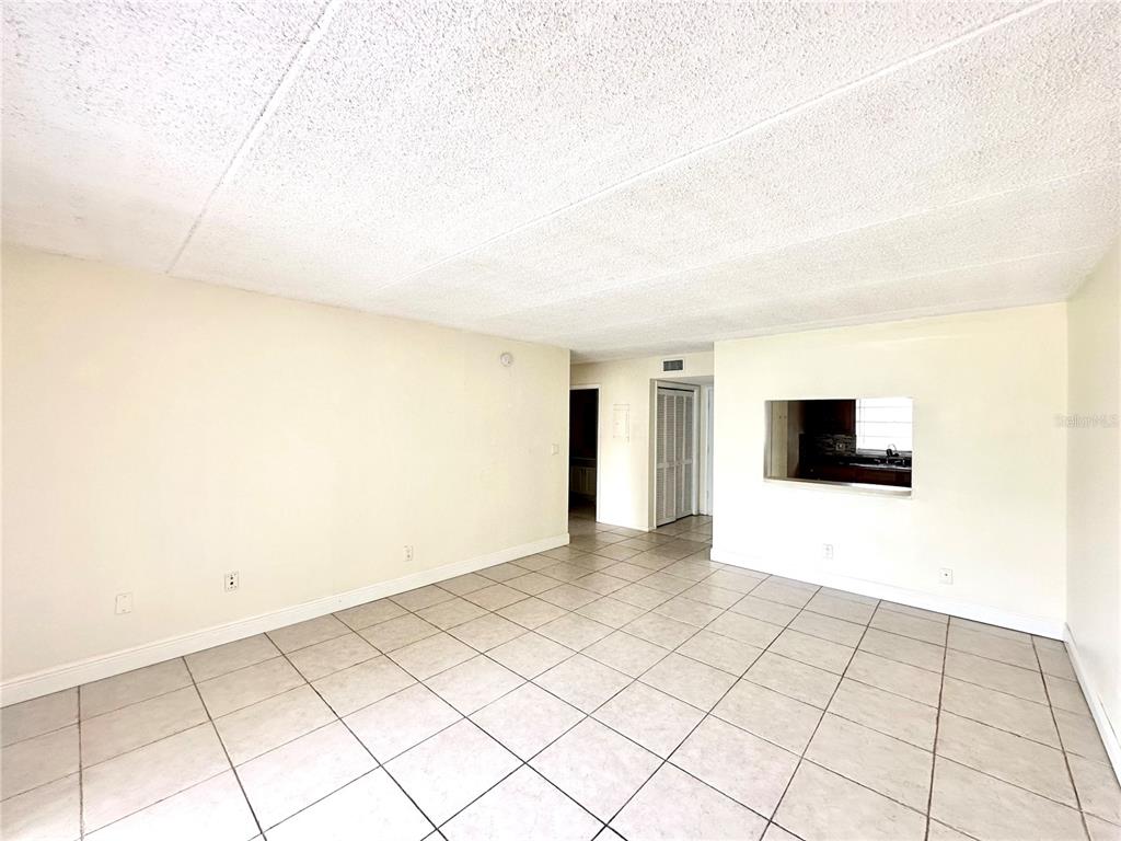 An empty living room with beige walls, light-colored tile flooring, and a kitchen pass-through window — 2830 SOMERSET PARK DRIVE UNIT 201, TAMPA, FL