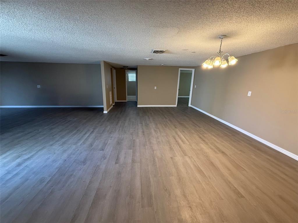 A spacious, empty room features light wood-look flooring, beige walls, and a central chandelier — 6104 SEABREEZE DRIVE, PORT RICHEY, FL