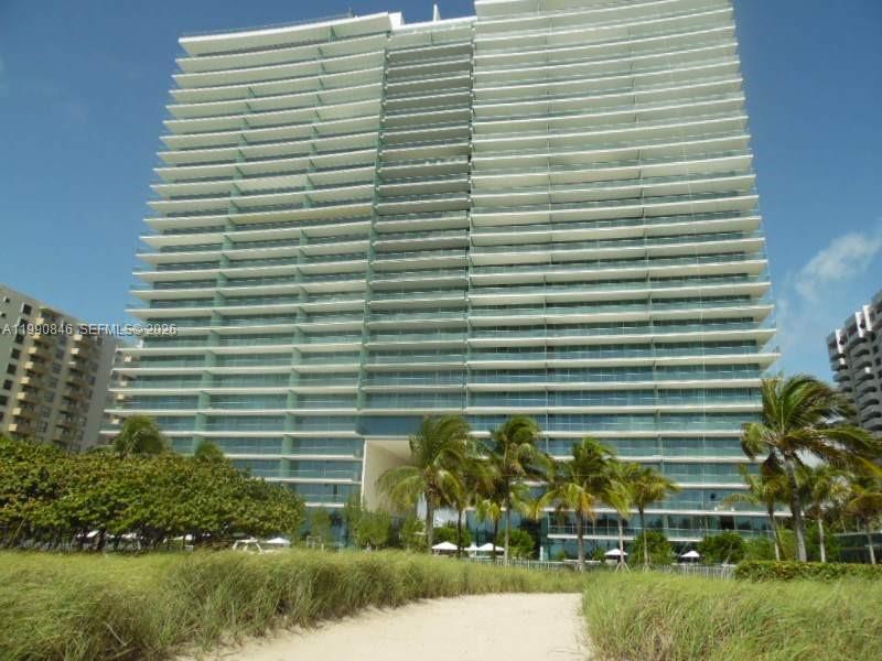 A tall, modern glass residential building with wrap-around balconies stands behind a sandy path — 10203 Collins Ave # 905, Bal Harbour FL 33154, BAL HARBOUR, FL