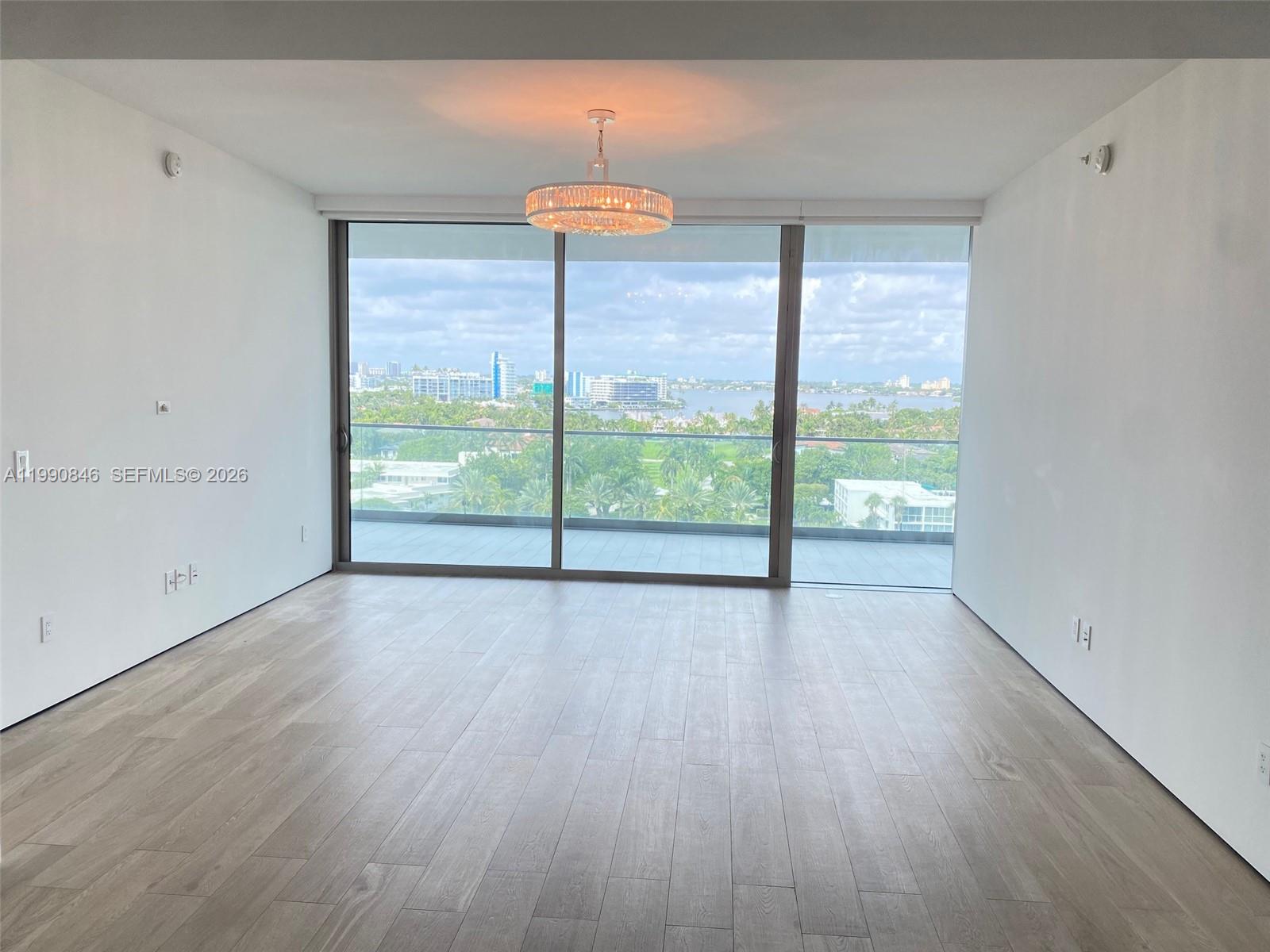 An empty room with light wood floors, a crystal chandelier, and floor-to-ceiling windows overlooking water — 10203 Collins Ave # 905, Bal Harbour FL 33154, BAL HARBOUR, FL