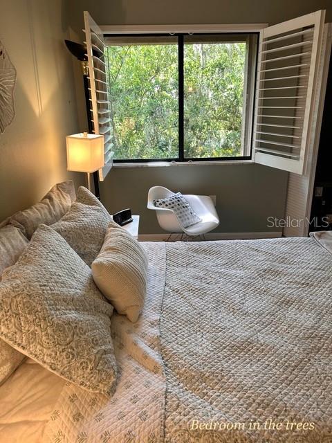 A bedroom features a bed with quilted bedding, a white accent chair, and window shutters — 1680 BROOKHOUSE CIRCLE UNIT BR209, SARASOTA, FL