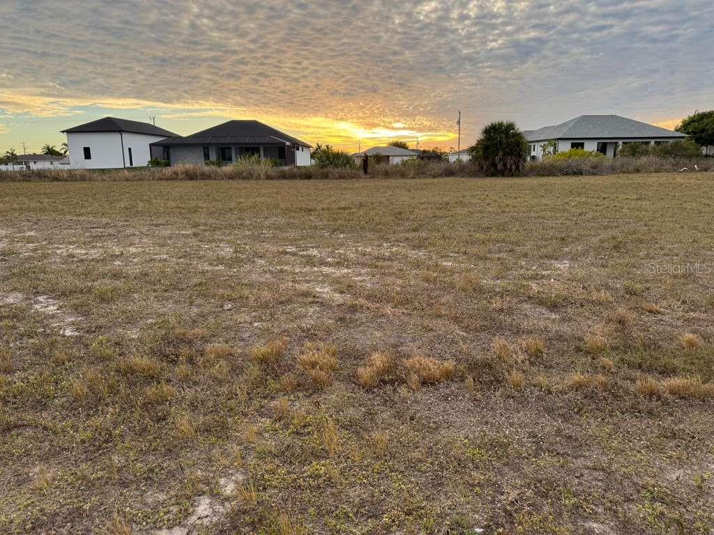 A vacant, grassy lot sits in front of several modern houses under a sunset sky — 1302 NE 19TH PLACE, CAPE CORAL, FL