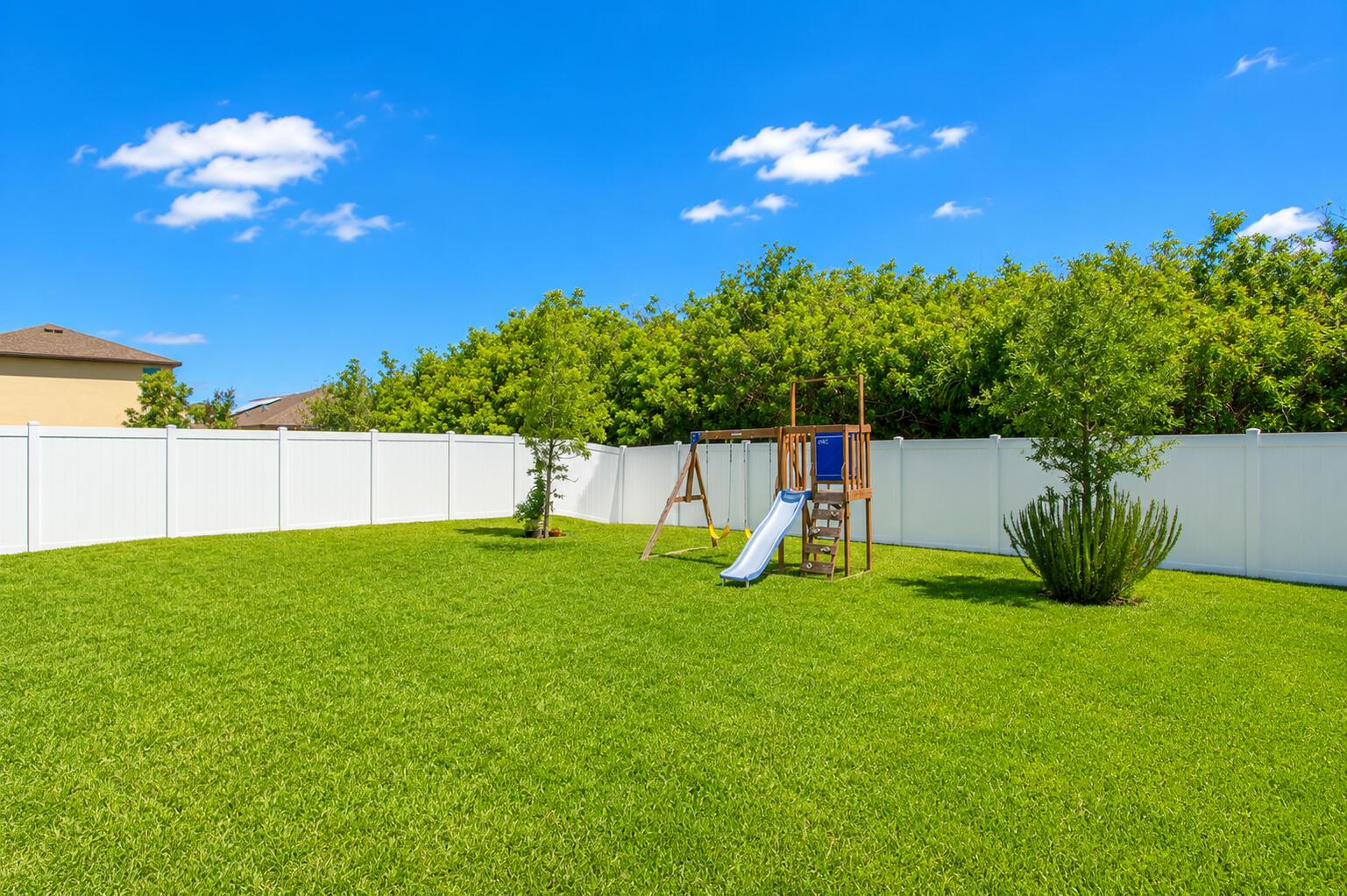 A grassy backyard features a wooden playset with a slide and a white vinyl fence — 4242 Thurso Way, Fort Pierce FL 34947, FORT PIERCE, FL