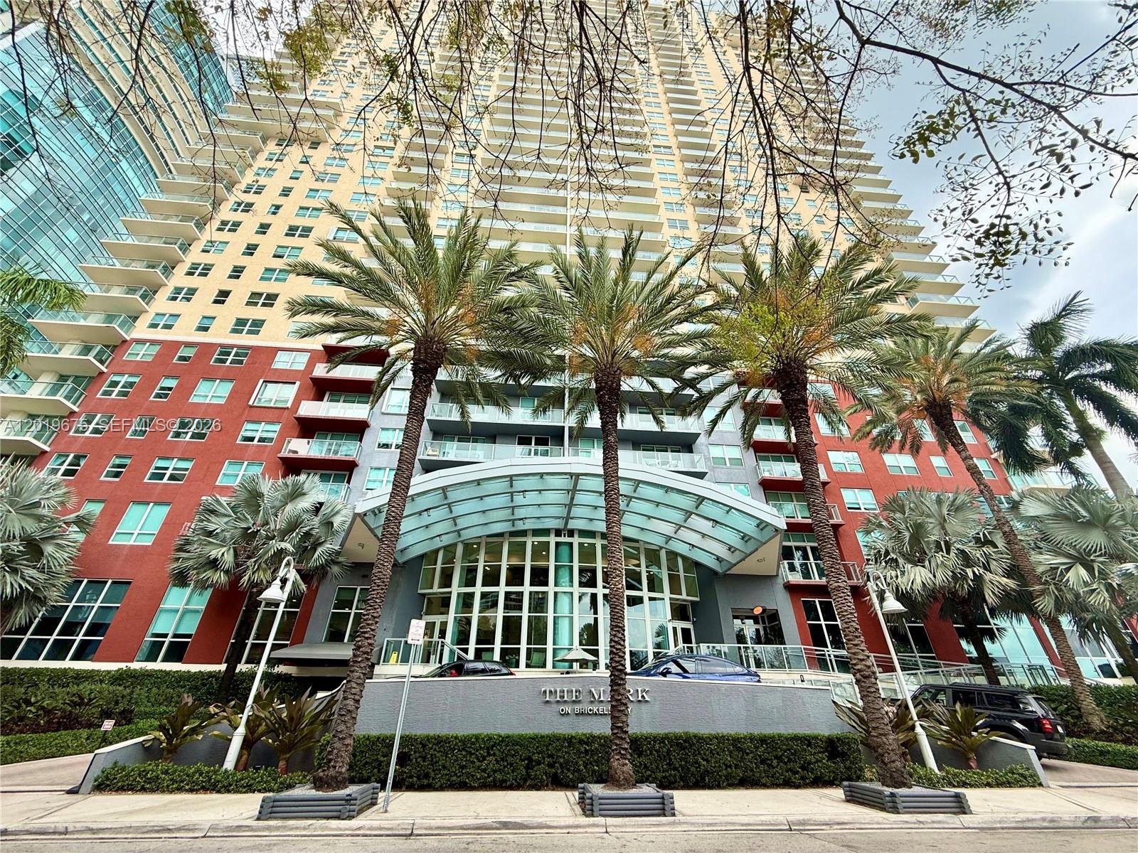 A low-angle view of a tall residential building with a glass entrance canopy and palm trees — 1155 Brickell Bay Dr # 1205, Miami FL 33131, MIAMI, FL